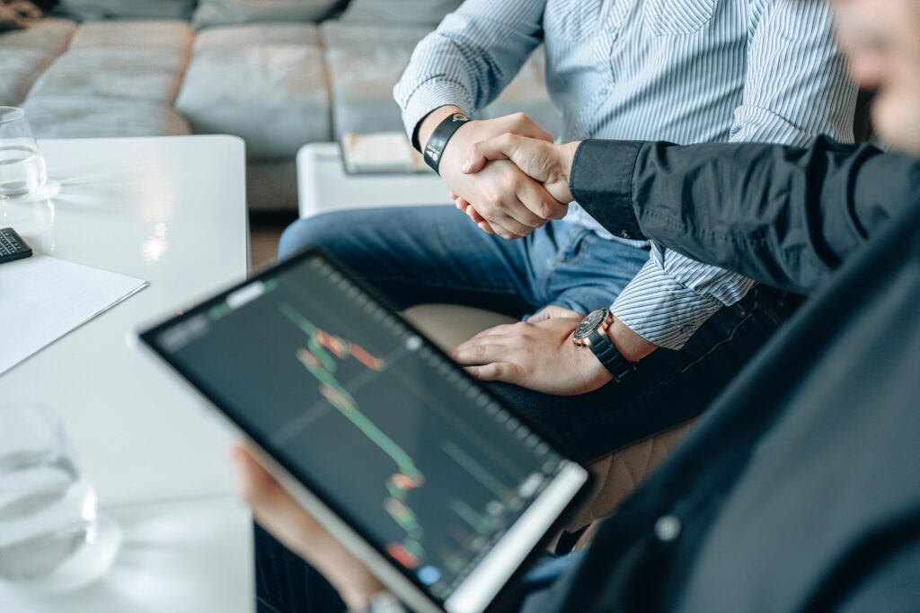 Home Two businesspeople shaking hands over stock market data on a tablet.